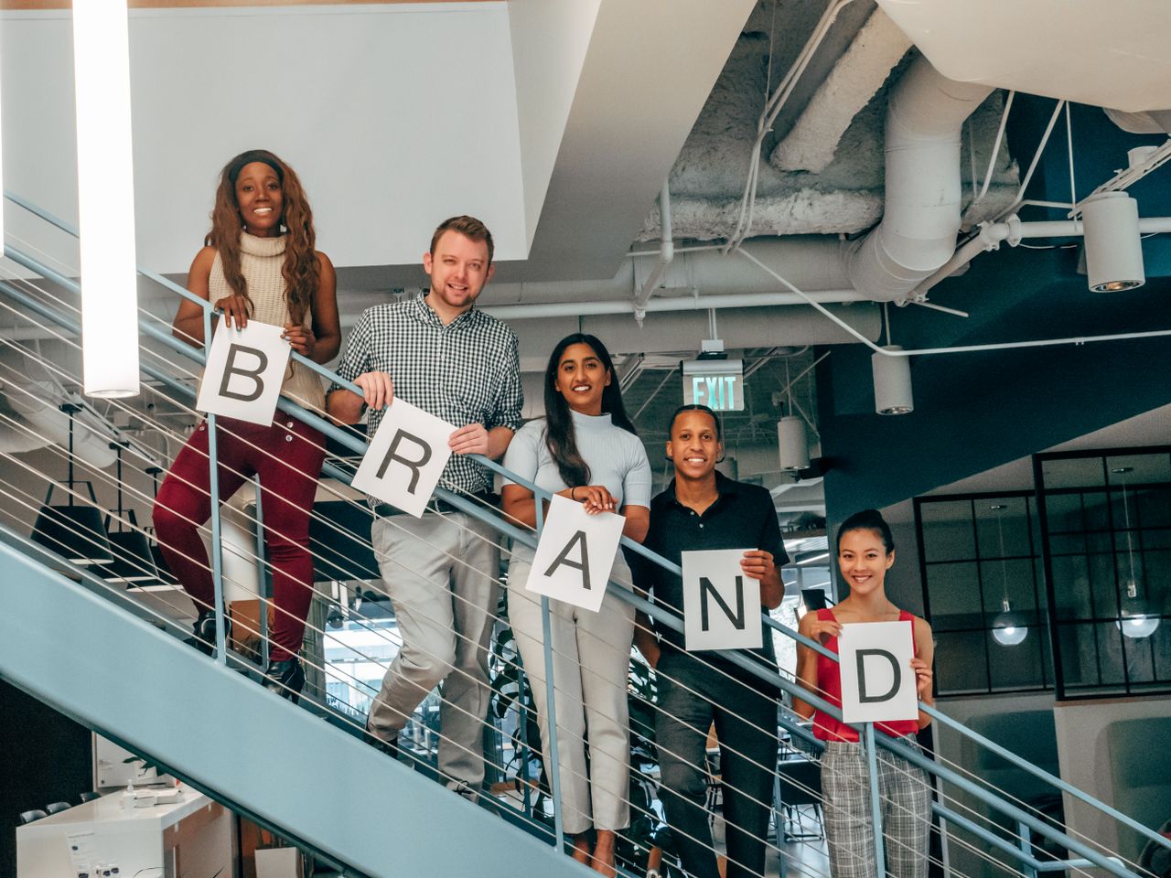 Diverse group of adults smiling and holding BRAND signs on an office staircase.