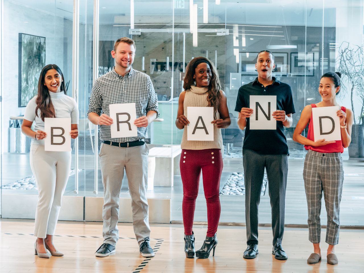 Diverse business team smiling and holding signs spelling BRAND in a modern office.